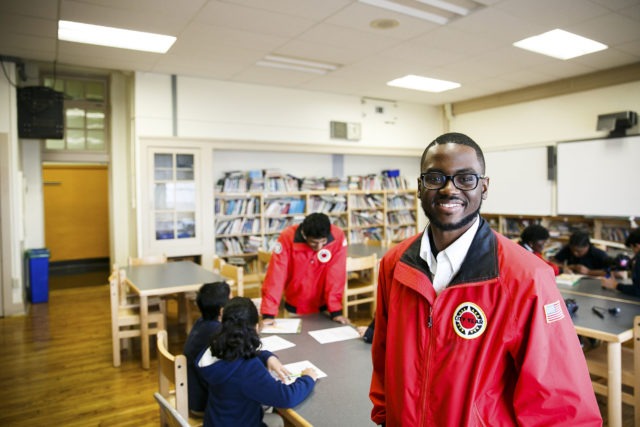 A ɫƵ AmeriCorps member stand in a library, looking proudly at the camera. Behind him are students working on homework with another AmeriCorps member