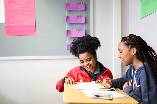 ɫƵ AmeriCorps member kneeling next to a student at a desk as they review work