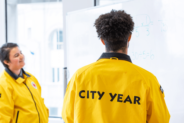 two ɫƵ AmeriCorps members in yellow jackets face a whiteboard with math problems on it